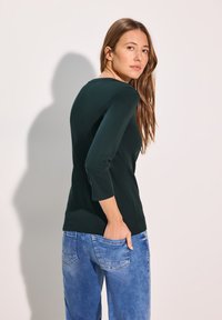 Musta vihreä istuva paita, jossa on kolme neljännesosaa hihat ja pyöreä kaula-aukko, yhdistettynä vaaleansinisiin farkkuihin, jotka korostavat rentoa istuvuutta.