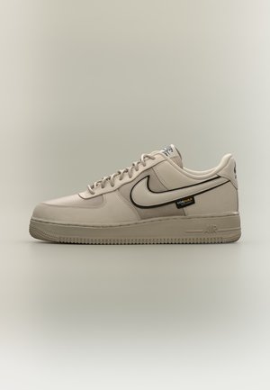 Baskets Nike Air Force 1 beiges avec swoosh bordé de noir, étiquette en tissu Cordura et semelle texturée sur fond neutre.