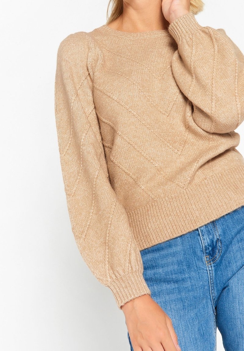 Beige stickad tröja med ett strukturerat diamantt mönster, rund halsringning och puffärmar, tillsammans med blå jeans.