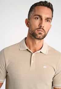 Camisa polo beige con tejido texturizado, que cuenta con un cuello clásico, tres botones y acentos blancos en contraste en el cuello y las mangas.
