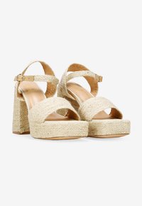Sandalias de esparto beige con textura trenzada, amplias correas cruzadas, tacón grueso y detalle de hebilla en el tobillo; suela de madera con plataforma.