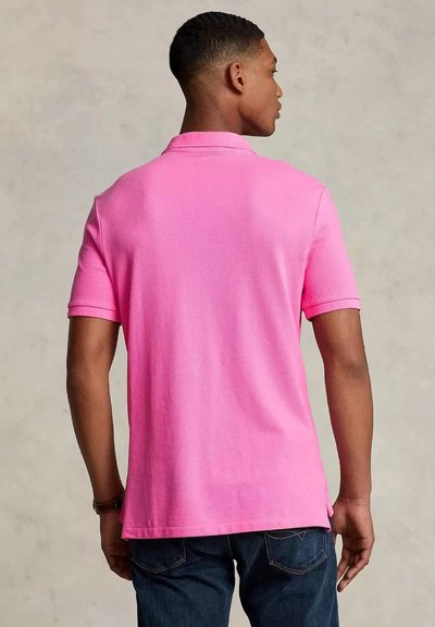 Un uomo sta di spalle rispetto all'osservatore, indossando una polo rosa brillante e jeans scuri, contro uno sfondo neutro.