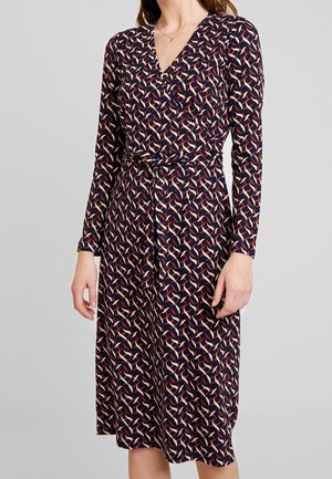 Frau trägt ein marineblaues Wickelkleid mit einem geometrischen Muster in Rot, Weiß und Beige und einem gebundenen Gürtel in der Taille, steht vor einem grauen Hintergrund.