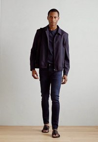 Malli, joka wear navy-takkia, tumman paidan, kapeita tummia farkkuja ja mustia sandaaleja, pitäen mustaa laukkua. Sujuva tekstuuri ja nykyaikainen muotoilu.