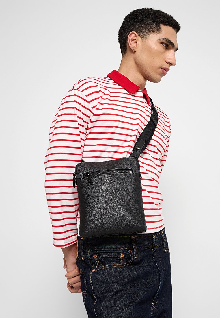 Sac en cuir noir avec une finition texturée, doté d'un compartiment zippé en haut et d'une poche avant. À porter en bandoulière avec une sangle réglable.