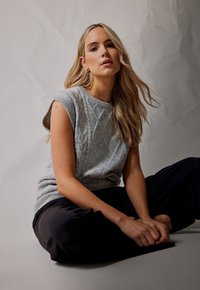 Maglione grigio lavorato a maglia senza maniche con motivi a diamante, abbinato a pantaloni neri. Modello seduto con le braccia incrociate sulle ginocchia, sfondo neutro.