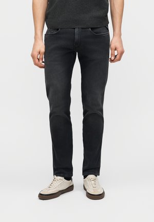 Άνδρας που φοράει στενά μαύρα τζιν και μπεζ αθλητικά παπούτσια, στέκεται μπροστά σε απλό φωτεινό φόντο.