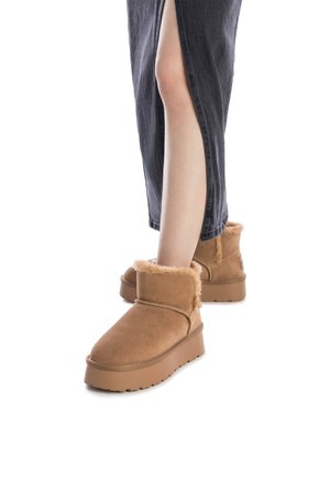 Botas de tobillo de ante marrón con forro de piel sintética, suela plana y detalles de costura lateral. Llevadas con una falda de mezclilla negra que presenta una abertura en el muslo.