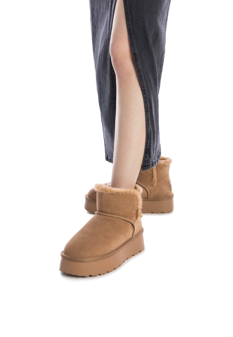 Botas de tobillo de ante marrón con forro de piel sintética, suela plana y detalles de costura lateral. Llevadas con una falda de mezclilla negra que presenta una abertura en el muslo.