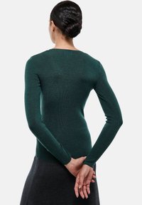 Pull en maille vert à manches longues avec un design ajusté, texture lisse et encolure ronde. Porté avec une jupe gris foncé, affichant un style minimaliste.