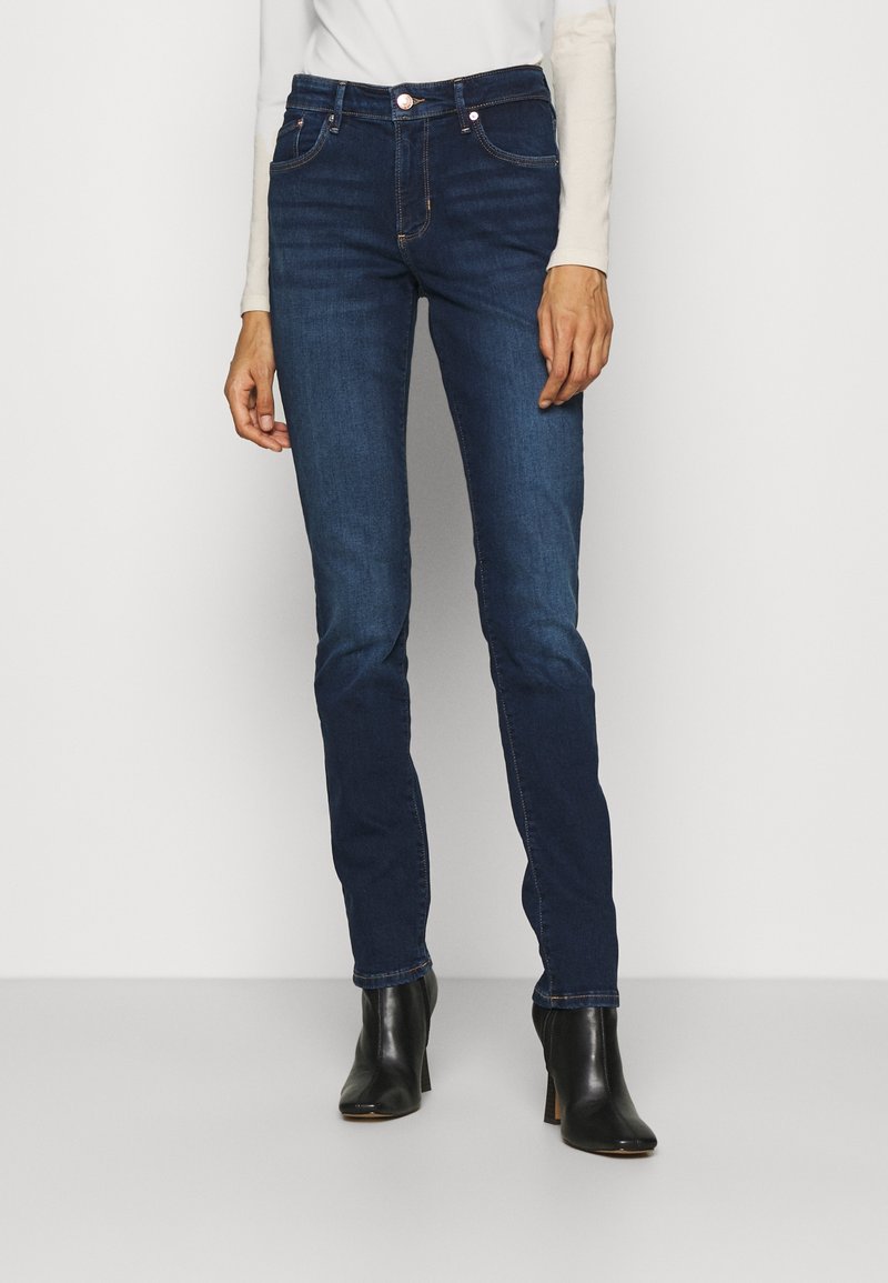 Jean slim bleu foncé porté avec des bottines noires à talons et un haut blanc à manches longues, visible de la taille jusqu'aux pieds.