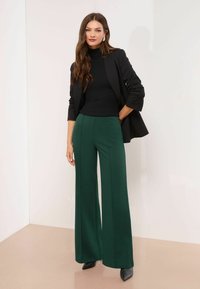 Mustaga soonik, rohelised laiad püksid, kohandatud must bleiser, sile tekstuur, mustad kontsad, seismine heleda taustaga.