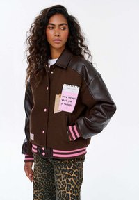 Kobieta z długimi kręconymi włosami nosi brązową kurtkę ze skórzanymi rękawami i różowo-pasiastymi mankietami, spodnie w lamparta i naszywkę z napisem "Rośnij przez to, co przechodzisz."