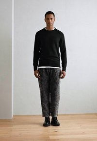 Suéter negro, capa blanca de cuello redondo debajo, pantalones rayados grises, zapatos negros pulidos, de pie sobre un suelo de madera contra un fondo neutro.