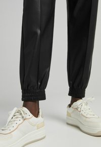 Schwarze Kunstlederhosen mit elastischen Bündchen, kombiniert mit weißen Sneakers mit beigen Akzenten und perforierten Bereichen.