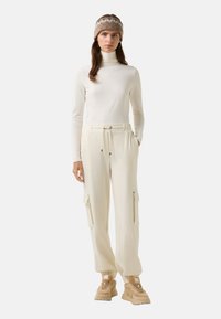 Crème turtle neck trui gecombineerd met crème cargo broek met ritszakken en een elastische tailleband. Model draagt beige sneakers en een patroondop.