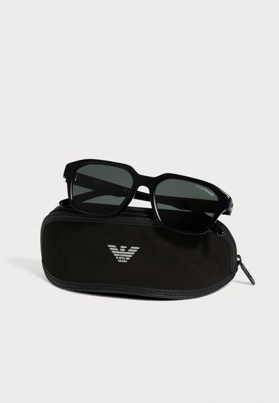Des lunettes de soleil noires Emporio Armani aux verres foncés reposent sur un étui rigide à fermeture éclair assorti, sur un fond clair.