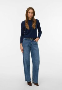 Sieviete stāv, valkājot tumši zilu džemperi, plašām biksēm ar melnu jostu un brūnus puszābakus uz vienkārša balta fona.