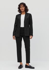 Twee-delige zwarte kostuum met een op maat gemaakte blazer met twee knopen en rechte broek. Gecombineerd met glanzende zwarte instappers.