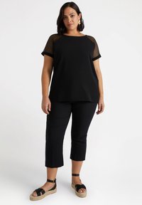 Zwarte top met mesh mouwen, gecombineerd met zwarte cropped broek. De outfit is voorzien van platte sandalen met een espadrille hak en enkelbandjes.