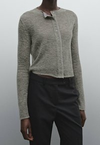 Henkilö, joka on pukeutunut vaaleanharmaaseen napitettuun lyhythihaiseen neuletakkiin ja tummiin korkeavyötäröisiin housuihin yksinkertaista harmaata taustaa vasten.