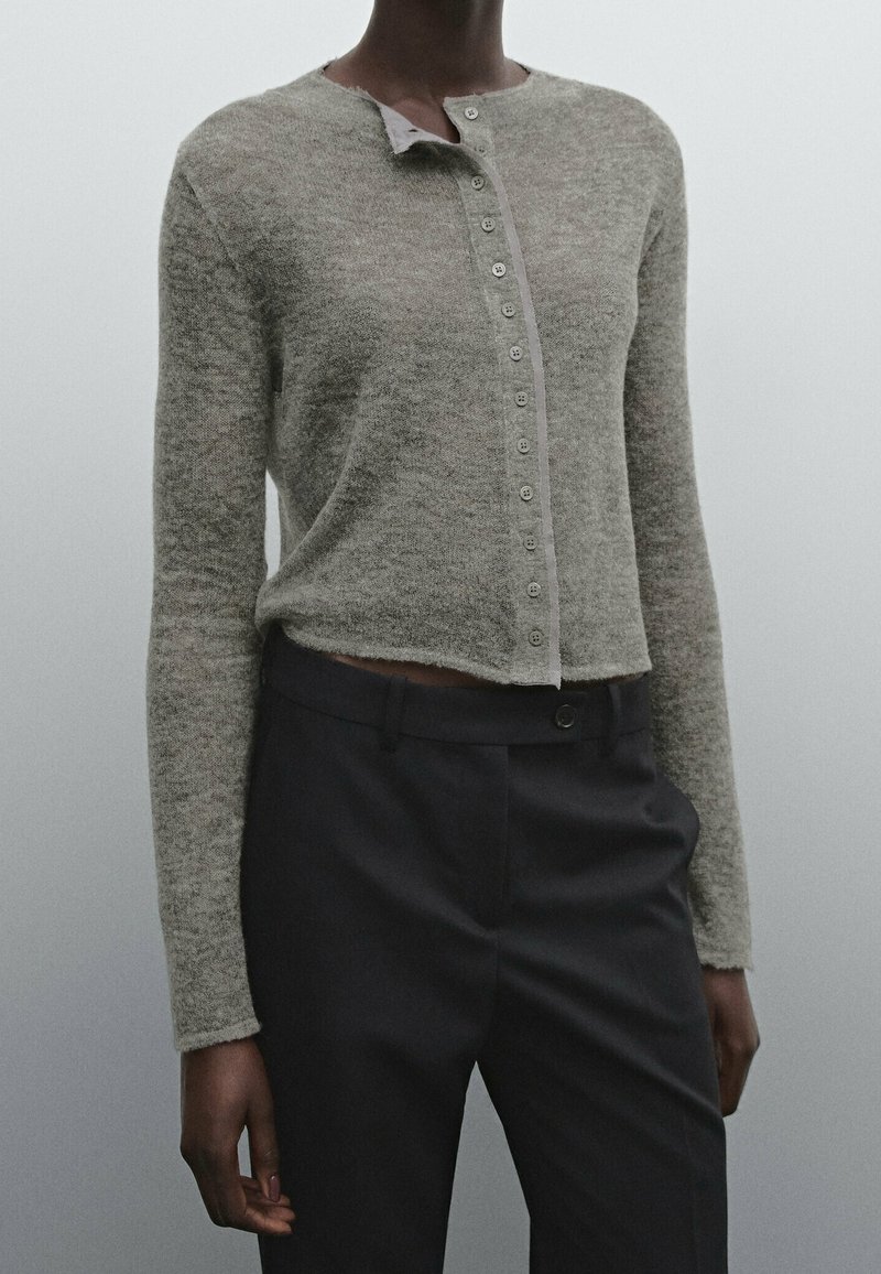 Henkilö, joka on pukeutunut vaaleanharmaaseen napitettuun lyhythihaiseen neuletakkiin ja tummiin korkeavyötäröisiin housuihin yksinkertaista harmaata taustaa vasten.