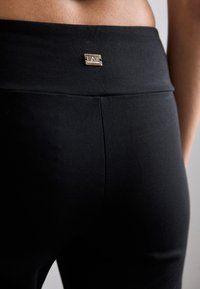 Black leggings with a smooth texture and a high waistband, featuring a small metallic logo detail on the back.