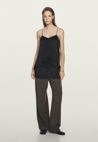 Camisola di satin nera con spalline sottili, sovrapposta a un pizzo nero, abbinata a pantaloni larghi di colore marrone scuro e scarpe nere.