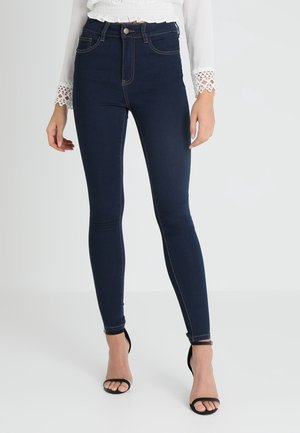 Kobieta ubrana w ciemnoniebieskie obcisłe jeansy, zestawione z czarnymi sandałami na wysokim obcasie oraz białą bluzką z długimi rękawami i koronkowymi mankietami.