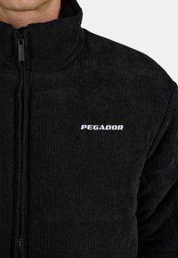 Sort corduroyjakke med høj krave, lynlåslukning og hvid "PEGADOR" logo på venstre bryst. Blød tekstur med ribbet mønster.