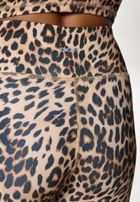 Legíny s leopardím vzorom v béžovej a čiernej farbe, s vysokým pásom a kontrastným prešitím. Logo značky viditeľné na páse.