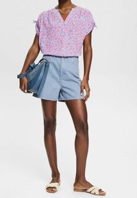 Lichtroze fleurige blouse met korte mouwen en een V-hals; gecombineerd met blauwe denim shorts en crèmekleurige sandalen. Houdt een blauwe handtas vast.