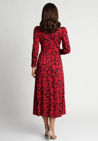 Kobieta stojąca tyłem, ubrana w czarną midi sukienkę w czerwony wzór róż oraz beżowe sandały na wysokim obcasie.