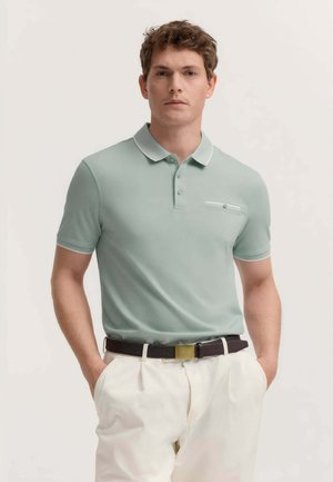 Uomo che indossa una polo verde chiaro a maniche corte con tasca sul petto, pantaloni bianchi e cintura nera con fibbia dorata, mani nelle tasche.