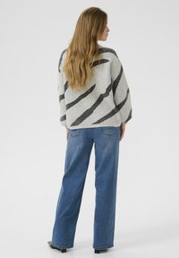 Femme aux longs cheveux portant un pull gris avec des rayures noires diagonales et un jean large bleu, se tenant de dos contre un fond blanc.
