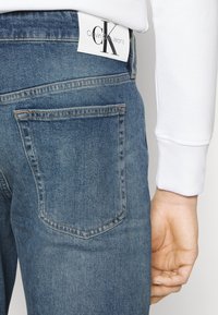 Modré džíny z denimu s lehkým praním, s zadní kapsou a bílou obdélníkovou visačkou se nápisem "Calvin Klein Jeans" v černé barvě.