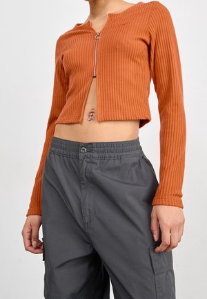 Torso met een cropped, geribd, verbrande oranje longsleeve met een rits aan de voorkant en donkergrijze cargobroek. Navelpiercing zichtbaar.