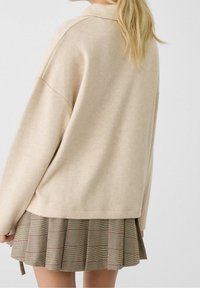 Maglione beige a maglia con una vestibilità rilassata e spalle scese, abbinato a una minigonna plissettata in tartan marrone con accenti rossi.