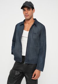 Tumši zila džinsa jaka ar teksturētu apdari, tai ir apkakle, divas priekšējās kabatas un rāvējslēdzēja aizdare. Valkāta virs baltas tanktopas.