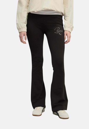 Persona que lleva pantalones negros acampanados con el logo de Puma en el muslo, zapatillas blancas y una chaqueta de color claro con puños elásticos.