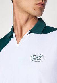 Camisola polo branca com detalhes em verde, apresentando um decote em V e mangas curtas. Logótipo EA7 notável no peito, tecido texturizado, design desportivo.