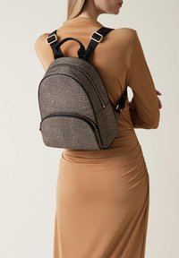 Schwarz-weiß gemusterter Rucksack mit zwei Reißverschlusstaschen, verstellbaren schwarzen Trägern und goldenen Hardware-Akzenten.