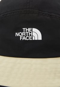 Cappello a secchi nero e beige in tessuto con il logo bianco de "The North Face" sul davanti, dotato di evidenti dettagli di cucitura.
