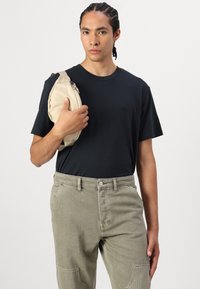 Svart t-shirt med en liten logotyp, olivgrön texturerad byxa med sidofickor och en rund beige väska som hålls under armen.