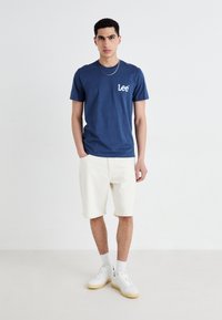 Tricou din bumbac bleumarin cu logo-ul "Lee" pe partea stângă a pieptului, asortat cu pantaloni scurți albi din denim și adidași albi. Guler simplu, rotund și mâneci scurte.