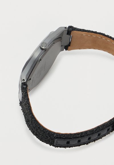 Ασημένιο μεταλλικό ρολόι χειρός με υφασμάτινο λουράκι μαύρου χρώματος με υφή και ορατή δερμάτινη εσωτερική πλευρά, εμφανιζόμενο από πλάγια και πίσω όψη.