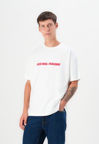 Hvit bomulls t-skjorte med rød tekst "RED BULL RACING", rund hals, korte ermer og løs passform. Bæres med blå denimjeans.