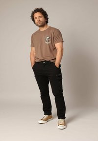 Bruine grafische t-shirt met bloemen schedelontwerp, zwarte cargobroeken met ritssluitingen, en lichtbruine sneakers met witte zolen.