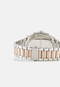 Reloj de acero inoxidable en plata y oro rosa con pulsera de eslabones, que presenta una caja redonda y detalles texturizados en la corona. Branding de Calvin Klein visible.