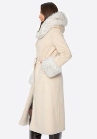 Beżowy wełniany płaszcz z kapturem i mankietami z sztucznego futra. Posiada pasek w talii, boczny rozcięcie oraz gładką fakturę. Minimalistyczny design, do kolan.